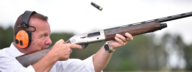 Photograph of sportsman wearing electronic hearing protection aiming and firing a shotgun with the spent shell casing of the previous shot is being ejected out of the ejection port.